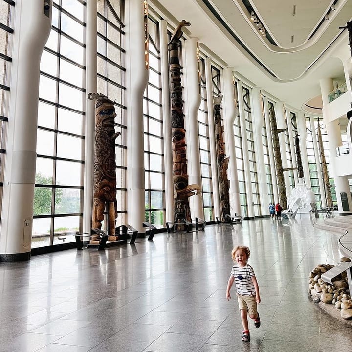 Blonde toddler running through a great hall with large windows, totem poles and a view of Canada’s parliament