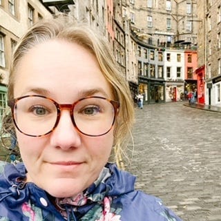 a blond female with glasses stands on a rainy cobble stone street in Edinburgh with colourful buildings in the background