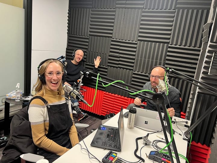 Three people sit in a room with large microphones in front of them. The walls are padded in black and red foam. Everyone is s