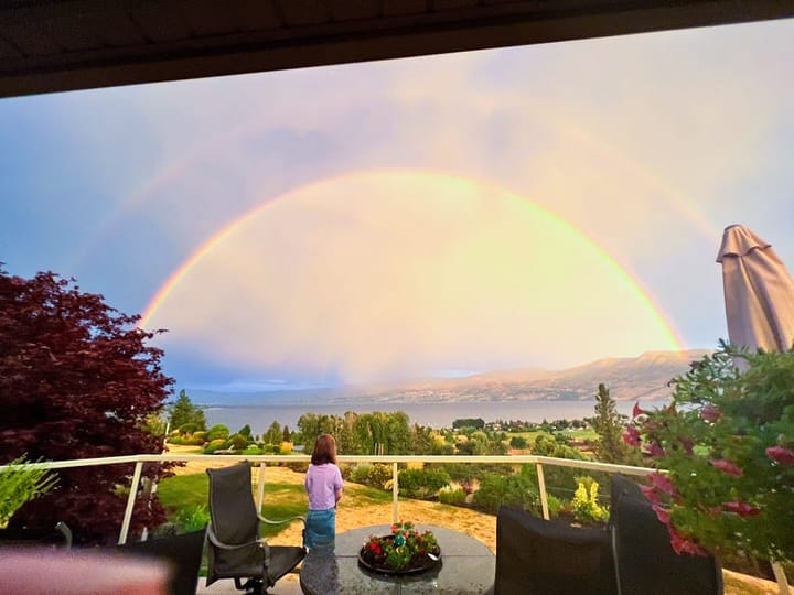 A young girl stands in front of a large double rainbow amidt storm clouds, overlooking a lake.