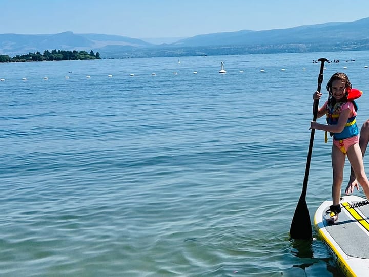 A young girl with a life jacket stands on a paddle board in a lake surrounded by mountains